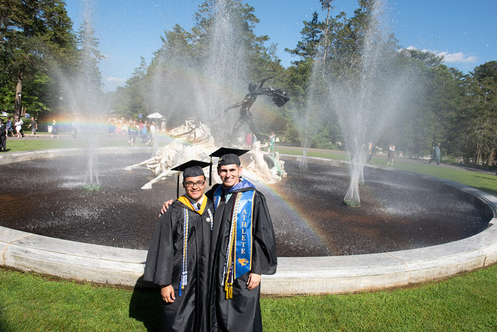 Two male graduates in cap and gown smiling in front of GCU apollo fountain.