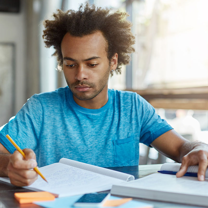 Handsome Afro American graduate student with curly hairstyle sitting at desk with book and copybook,