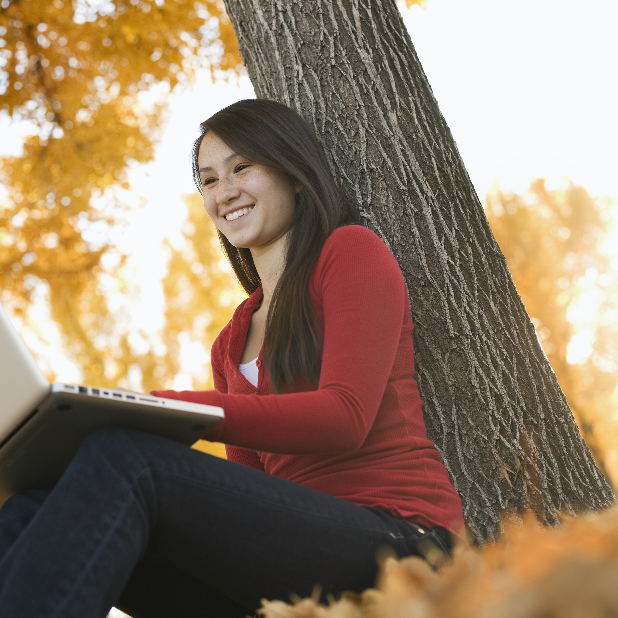 A young woman seated by a tree using a laptop computer.