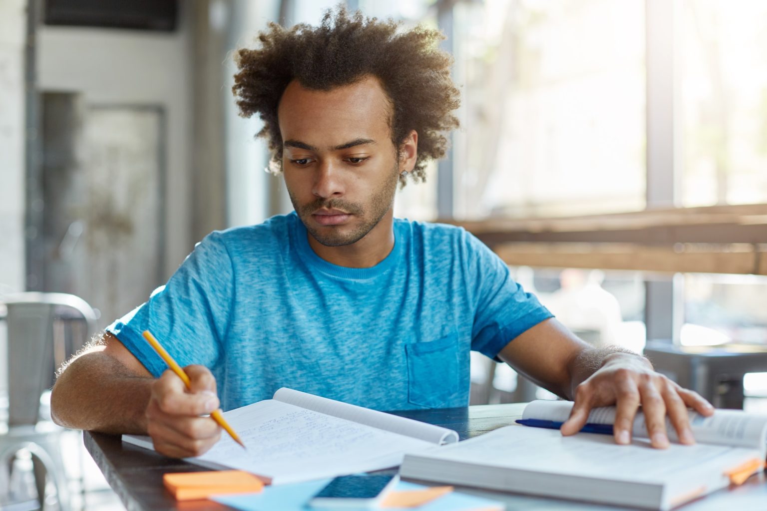 Handsome Afro American graduate student with curly hairstyle sitting at desk with book and copybook,
