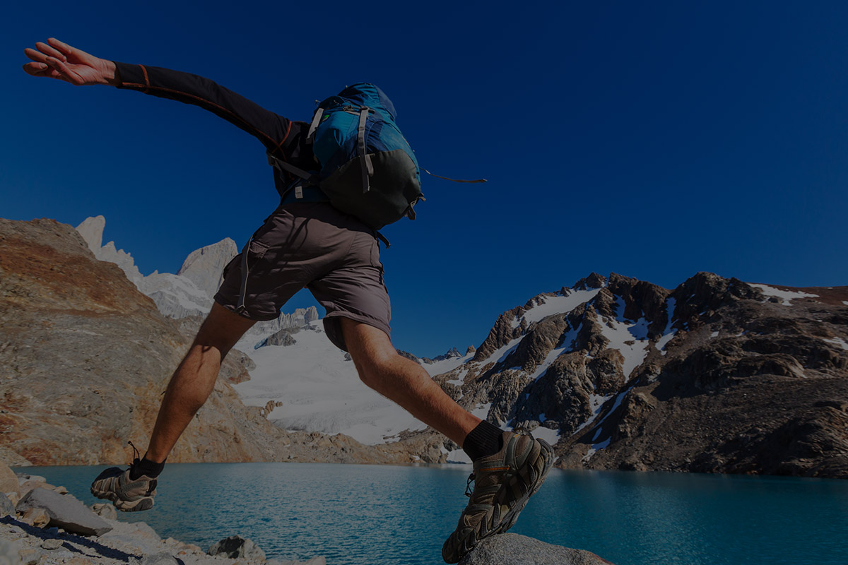 Hiker jumping across rocks in front of lake and mountains with blue sky.